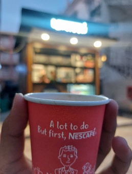 A hand is holding a red paper cup that has the phrase 'A lot to do, But first, NESCAF&Eacute;' printed on it. In the background, the image is blurred but shows a coffee kiosk with glowing lights at dusk.