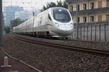 A modern high-speed train travels along railway tracks. The train is sleek, with a white exterior and blue accents. It moves through an urban area, passing alongside a building and overhead power lines. The tracks are surrounded by gravel and metal structures, with city buildings visible in the background.