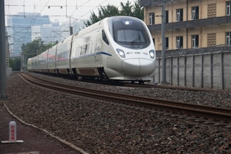 A modern high-speed train travels along railway tracks. The train is sleek, with a white exterior and blue accents. It moves through an urban area, passing alongside a building and overhead power lines. The tracks are surrounded by gravel and metal structures, with city buildings visible in the background.