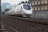A modern high-speed train travels along railway tracks. The train is sleek, with a white exterior and blue accents. It moves through an urban area, passing alongside a building and overhead power lines. The tracks are surrounded by gravel and metal structures, with city buildings visible in the background.