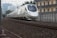 A modern high-speed train travels along railway tracks. The train is sleek, with a white exterior and blue accents. It moves through an urban area, passing alongside a building and overhead power lines. The tracks are surrounded by gravel and metal structures, with city buildings visible in the background.