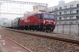 A red locomotive train is traveling on railway tracks with several carriages behind it. The train tracks run parallel to a modern multi-story apartment building on the right side of the image. Overhead electrical wires and support structures are visible above the train, indicating an electrified railway. The area has an urban setting with buildings and infrastructure in the background. The ground beside the tracks is covered with gravel and some red dirt patches.