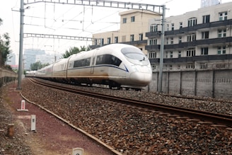 A sleek, modern high-speed train travels along a railway track beside urban residential buildings. Overhead electric power lines stretch across the scene, and the railway is surrounded by a gravel bed. The train's aerodynamic design and large windows are prominent, indicating its capacity for fast travel.