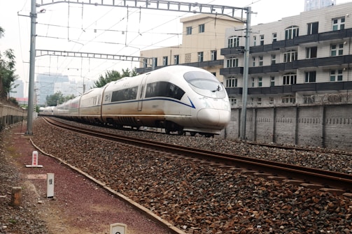 A sleek, modern high-speed train travels along a railway track beside urban residential buildings. Overhead electric power lines stretch across the scene, and the railway is surrounded by a gravel bed. The train's aerodynamic design and large windows are prominent, indicating its capacity for fast travel.