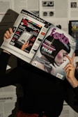 A person holds two magazines in front of their face, obscuring their identity. The left magazine features technology-focused content with a headline for 'Digital Vision' and an image of an e-reader. The right magazine, titled 'foto', shows a close-up of a woman with a short, dark hairstyle. The background consists of more magazine pages or printed text. The lighting casts shadows, creating a dramatic effect.