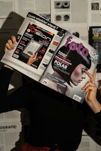 A person holds two magazines in front of their face, obscuring their identity. The left magazine features technology-focused content with a headline for 'Digital Vision' and an image of an e-reader. The right magazine, titled 'foto', shows a close-up of a woman with a short, dark hairstyle. The background consists of more magazine pages or printed text. The lighting casts shadows, creating a dramatic effect.