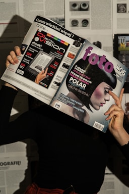 A person holds two magazines in front of their face, obscuring their identity. The left magazine features technology-focused content with a headline for 'Digital Vision' and an image of an e-reader. The right magazine, titled 'foto', shows a close-up of a woman with a short, dark hairstyle. The background consists of more magazine pages or printed text. The lighting casts shadows, creating a dramatic effect.
