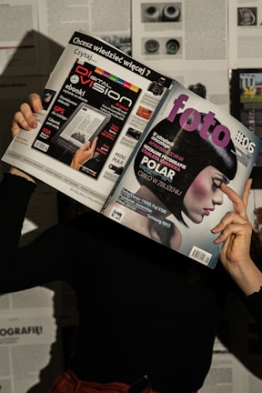 A person holds two magazines in front of their face, obscuring their identity. The left magazine features technology-focused content with a headline for 'Digital Vision' and an image of an e-reader. The right magazine, titled 'foto', shows a close-up of a woman with a short, dark hairstyle. The background consists of more magazine pages or printed text. The lighting casts shadows, creating a dramatic effect.