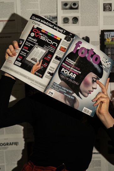 A person holds two magazines in front of their face, obscuring their identity. The left magazine features technology-focused content with a headline for 'Digital Vision' and an image of an e-reader. The right magazine, titled 'foto', shows a close-up of a woman with a short, dark hairstyle. The background consists of more magazine pages or printed text. The lighting casts shadows, creating a dramatic effect.