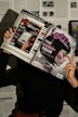 A person holds two magazines in front of their face, obscuring their identity. The left magazine features technology-focused content with a headline for 'Digital Vision' and an image of an e-reader. The right magazine, titled 'foto', shows a close-up of a woman with a short, dark hairstyle. The background consists of more magazine pages or printed text. The lighting casts shadows, creating a dramatic effect.