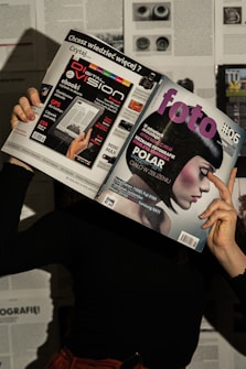 A person holds two magazines in front of their face, obscuring their identity. The left magazine features technology-focused content with a headline for 'Digital Vision' and an image of an e-reader. The right magazine, titled 'foto', shows a close-up of a woman with a short, dark hairstyle. The background consists of more magazine pages or printed text. The lighting casts shadows, creating a dramatic effect.