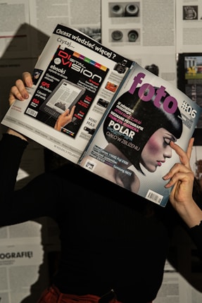 A person holds two magazines in front of their face, obscuring their identity. The left magazine features technology-focused content with a headline for 'Digital Vision' and an image of an e-reader. The right magazine, titled 'foto', shows a close-up of a woman with a short, dark hairstyle. The background consists of more magazine pages or printed text. The lighting casts shadows, creating a dramatic effect.