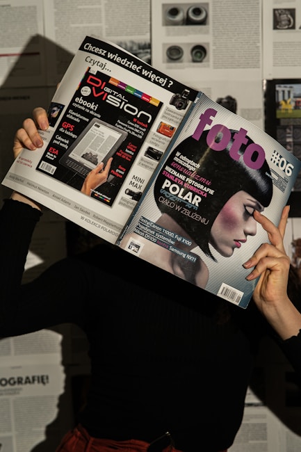 A person holds two magazines in front of their face, obscuring their identity. The left magazine features technology-focused content with a headline for 'Digital Vision' and an image of an e-reader. The right magazine, titled 'foto', shows a close-up of a woman with a short, dark hairstyle. The background consists of more magazine pages or printed text. The lighting casts shadows, creating a dramatic effect.