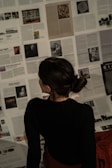 A person with dark hair is sitting on a floor covered with scattered pages from magazines or newspapers, appearing to be deeply engaged in reading or examining the content. The text and images on the pages vary, creating a mosaic-like background.