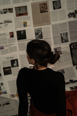 A person with dark hair is sitting on a floor covered with scattered pages from magazines or newspapers, appearing to be deeply engaged in reading or examining the content. The text and images on the pages vary, creating a mosaic-like background.