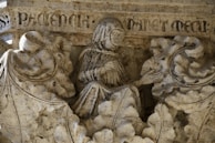 Detailed stone carving depicting a robed figure surrounded by ornate foliage, with inscriptions in Latin at the top. The intricacy of the foliage contrasts with the serene expression of the central figure.