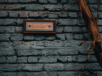 A textured brick wall painted in a dark teal color with a framed decorative script hanging on it. The frame contains intricate calligraphy surrounded by ornate artistic designs in a contrasting color. A dry vine or branch runs vertically across the right side of the wall, adding a natural, rustic element to the scene.