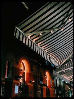 An outdoor scene featuring a series of striped awnings illuminated by string lights, creating a cozy and inviting atmosphere. The brick building beneath the awnings has a series of arched windows, and warm light emanates from within.