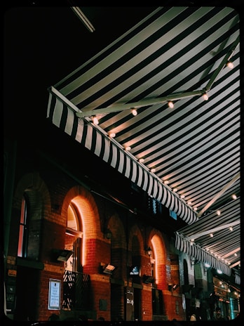 An outdoor scene featuring a series of striped awnings illuminated by string lights, creating a cozy and inviting atmosphere. The brick building beneath the awnings has a series of arched windows, and warm light emanates from within.