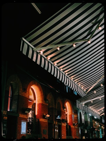 An outdoor scene featuring a series of striped awnings illuminated by string lights, creating a cozy and inviting atmosphere. The brick building beneath the awnings has a series of arched windows, and warm light emanates from within.