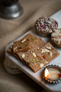 A close-up view of pieces of nut-studded sweets arranged on a white marble slab. Next to the sweets, there are two small bowls, one containing a mix of colorful seeds and the other filled with almond slivers. A lit diya (oil lamp) is placed near the sweets, adding a warm glow to the scene.