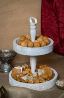 A two-tiered white tray holds an assortment of Indian sweets. The top tier is filled with round brown ladoos, while the bottom tier contains squares of Indian barfi, some covered with silver leaf. The tray is set against a textured brown background with a red fabric draped to one side, and additional serving bowls are partially visible.