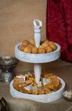 A two-tiered white tray holds an assortment of Indian sweets. The top tier is filled with round brown ladoos, while the bottom tier contains squares of Indian barfi, some covered with silver leaf. The tray is set against a textured brown background with a red fabric draped to one side, and additional serving bowls are partially visible.