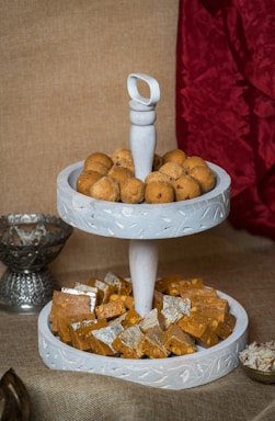A two-tiered white tray holds an assortment of Indian sweets. The top tier is filled with round brown ladoos, while the bottom tier contains squares of Indian barfi, some covered with silver leaf. The tray is set against a textured brown background with a red fabric draped to one side, and additional serving bowls are partially visible.