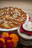 A tantalizing dessert displayed on a white plate, garnished with slivered almonds and pistachios. The setting includes festive decorations, with marigold flowers and rose petals nearby. A small bowl with a similar dessert or dip is placed on the side, on top of a decorative stand.