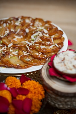 A tantalizing dessert displayed on a white plate, garnished with slivered almonds and pistachios. The setting includes festive decorations, with marigold flowers and rose petals nearby. A small bowl with a similar dessert or dip is placed on the side, on top of a decorative stand.
