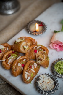 A series of intricately styled pastries garnished with almond slivers and rose petals, presented on a white marble slab. A small lit candle in an ornate holder and a pink rose are placed beside the pastries, adding a festive and elegant touch. Decorative bowls containing crushed nuts and green garnish accompany the setup.