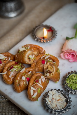 A series of intricately styled pastries garnished with almond slivers and rose petals, presented on a white marble slab. A small lit candle in an ornate holder and a pink rose are placed beside the pastries, adding a festive and elegant touch. Decorative bowls containing crushed nuts and green garnish accompany the setup.