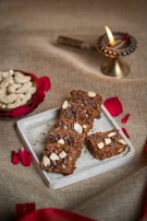 A set of four brown nut-topped squares are arranged on a small rectangular tray. They are surrounded by red rose petals and a small bowl filled with cashew nuts. In the background, a traditional oil lamp is lit, adding a warm glow. The setting is on a textured fabric surface.