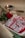 A traditional Indian sweet decorated with silver foil and rose petals is placed on a rectangular tray. Nearby, a lit brass oil lamp and a red rose enhance the aesthetic with warm lighting, all set against a textured, neutral-colored tablecloth.