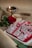 A traditional Indian sweet decorated with silver foil and rose petals is placed on a rectangular tray. Nearby, a lit brass oil lamp and a red rose enhance the aesthetic with warm lighting, all set against a textured, neutral-colored tablecloth.