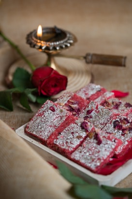A traditional Indian sweet decorated with silver foil and rose petals is placed on a rectangular tray. Nearby, a lit brass oil lamp and a red rose enhance the aesthetic with warm lighting, all set against a textured, neutral-colored tablecloth.