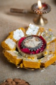 A plate of traditional Indian sweets adorned with edible silver foil and almond slices, placed around a decorative bowl containing a candle surrounded by rose petals. In the background, a lit brass oil lamp adds a warm, festive atmosphere.