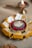 A plate of traditional Indian sweets adorned with edible silver foil and almond slices, placed around a decorative bowl containing a candle surrounded by rose petals. In the background, a lit brass oil lamp adds a warm, festive atmosphere.