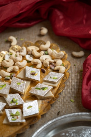 An arrangement of diamond-shaped silver-topped sweets garnished with sliced pistachios is presented on a decorative yellow plate. Surrounding the sweets are scattered cashew nuts. The plate is placed on a textured surface with red fabric softly draping in the background.