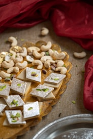 An arrangement of diamond-shaped silver-topped sweets garnished with sliced pistachios is presented on a decorative yellow plate. Surrounding the sweets are scattered cashew nuts. The plate is placed on a textured surface with red fabric softly draping in the background.