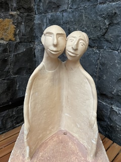 A clay sculpture depicting two abstract human figures joined together. Both figures are stylized with simple, exaggerated facial features and are color tones of beige and light brown. The background consists of a textured, dark stone wall, creating a contrasting background.