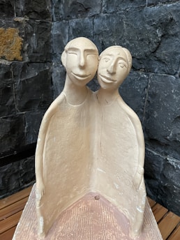 A clay sculpture depicting two abstract human figures joined together. Both figures are stylized with simple, exaggerated facial features and are color tones of beige and light brown. The background consists of a textured, dark stone wall, creating a contrasting background.