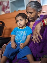 An elderly woman with silver hair, wearing a purple sari and colorful bangles, is holding a young child on her lap. The child is wearing a bright blue t-shirt and shorts. They are positioned indoors against a wooden background, with the woman appearing to look at the child fondly.