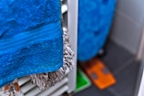 A close-up of a blue towel hanging on a radiator, with a fuzzy mop or cleaning cloth visible behind it. The floor has dark tiles, and a small orange and green object can be seen in the corner.