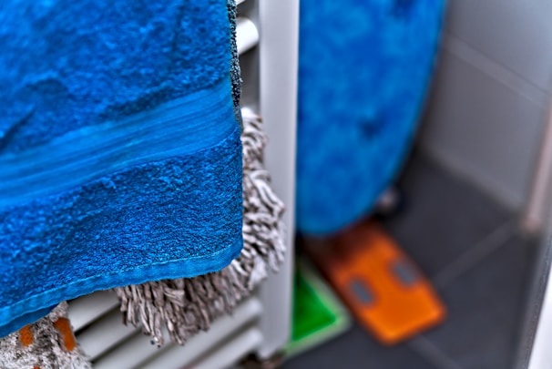 A close-up of a blue towel hanging on a radiator, with a fuzzy mop or cleaning cloth visible behind it. The floor has dark tiles, and a small orange and green object can be seen in the corner.