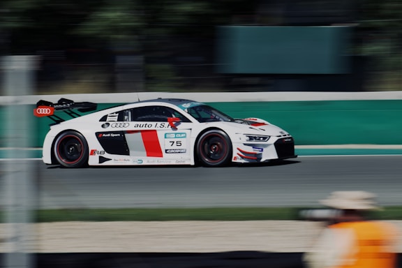 A sleek, white and red racing car speeds down a track, featuring prominent branding and logo placements on the side. The background is blurred, emphasizing the fast motion of the vehicle. A person wearing a beige hat and orange vest is visible in the foreground, indicating a possible race official or photographer.