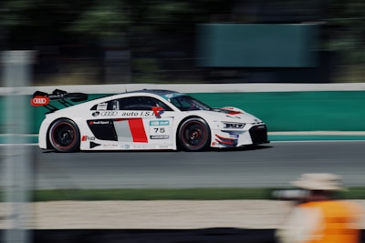 A sleek, white and red racing car speeds down a track, featuring prominent branding and logo placements on the side. The background is blurred, emphasizing the fast motion of the vehicle. A person wearing a beige hat and orange vest is visible in the foreground, indicating a possible race official or photographer.