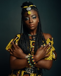 A woman with long, braided hair adorned with beads and threaded decorations stands with her arms crossed over her chest. She wears a vibrant yellow and brown patterned top and a colorful beaded bracelet on one wrist. Her makeup is bold, including blue eyeshadow and glossy lips. The background is a dark, neutral color, emphasizing her pose and attire.