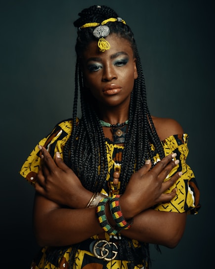 A woman with long, braided hair adorned with beads and threaded decorations stands with her arms crossed over her chest. She wears a vibrant yellow and brown patterned top and a colorful beaded bracelet on one wrist. Her makeup is bold, including blue eyeshadow and glossy lips. The background is a dark, neutral color, emphasizing her pose and attire.
