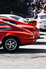 A line of high-performance sports cars, predominantly in red and white colors, parked in an outdoor setting. People can be seen in the background, suggesting an event or car show atmosphere.
